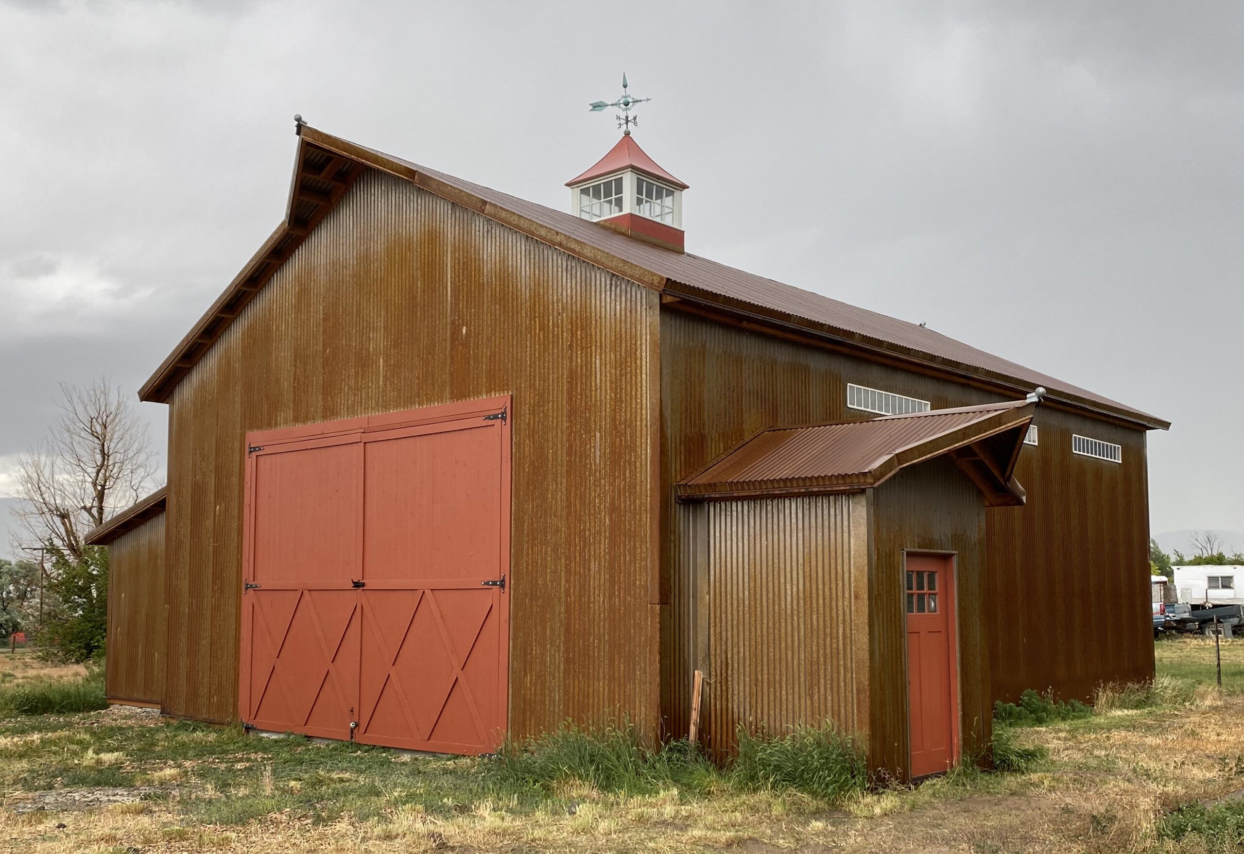 A large rustic barn with a corrugated metal exterior and red doors, featuring a small cupola on the roof with a weather vane on top.